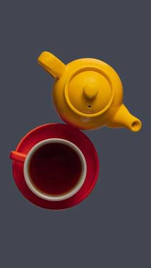 Overhead view of a yellow teapot and red tea cup with a saucer on dark gray background.