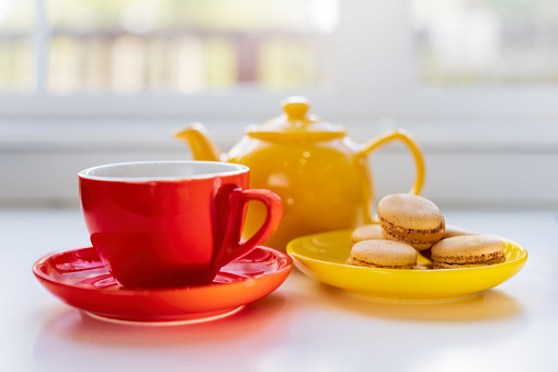 Breakfast tea in a red cup with macaroons served on yellow saucer with a yellow teapot in the background
