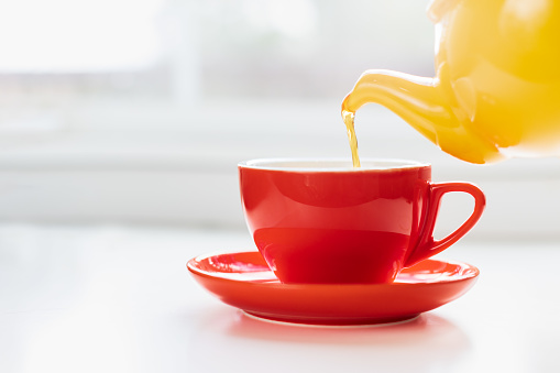 Red cup of tea being poured from a yellow teapot. Natural light freezes tea stream