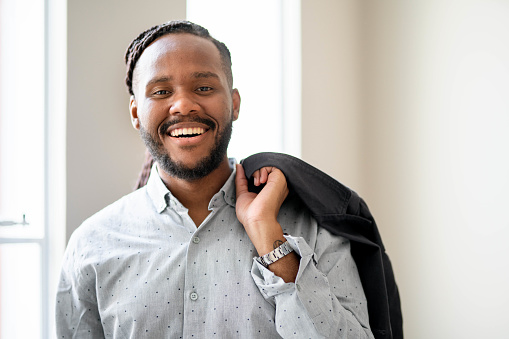 Portrait of a young businessman laughing while standing in an bright office with his blazer draped over his shoulder