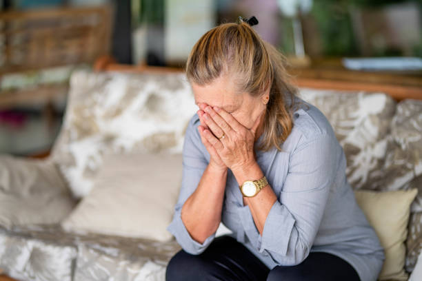 Senior woman with depression sitting with her head in her hands at home Depressed senior woman sitting alone with her head in her hands on a living room sofa at home older woman eyes closed stock pictures, royalty-free photos & images