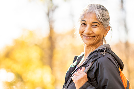 A senior woman of Middle Eastern decent is seen hiking alone on a sunny fall day.  She is dressed comfortably in athletic clothing and wearing a backpack as she smiles gently.