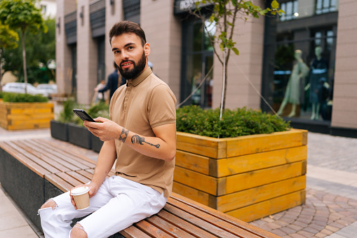 Portrait of brutal bearded young man holding using smartphone sitting on bench with takeaway coffee looking at camera on urban street. Tattooed male hatting on social media on summer day.
