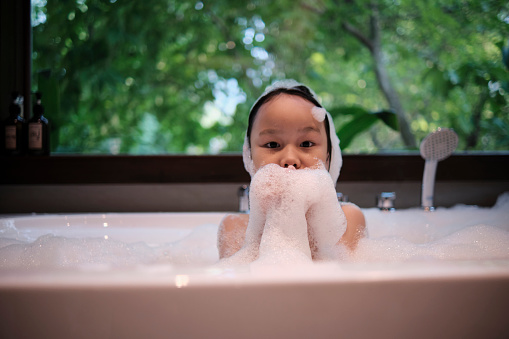 Portrait of one Asian little cute girl with happiness and fun playing with body soap foam, shampoo splash, and water bubbles in a white bathtub at a hotel bathroom with green tree window background.