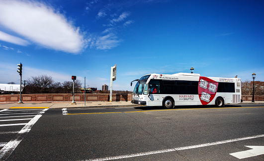 Cambridge, Massachusetts, USA - March 31, 2023:   A white Harvard University Shuttle Bus traveling from Cambridge to Allston (Boston) across the Anderson Memorial Bridge over the Charles River. The Harvard Veritas logo is on the side of the bus. The Harvard Shuttle buses serve the Cambridge and Allston (Boston) campuses.
