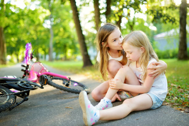 Adorable girl comforting her little sister after she fell off her bike at summer park. Child getting hurt while riding a bicycle. stock photo
