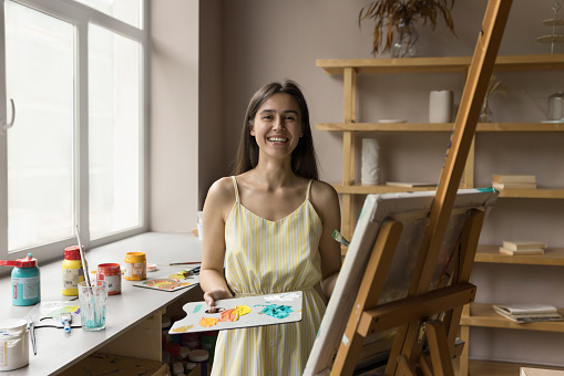 Happy beautiful young art school student drawing on canvas at easel, using artistic supplies, holding palette with acrylic, oil paints, looking at camera, smiling. Craft professional portrait