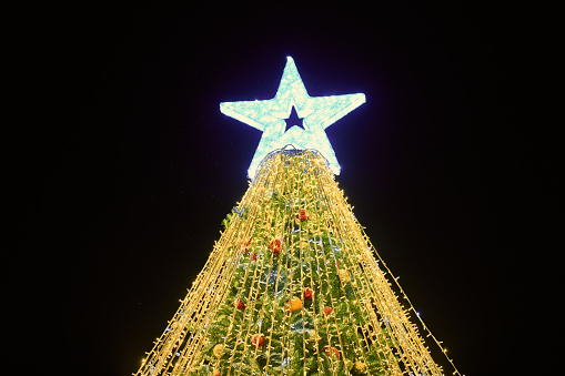 Christmas tree with yellow garlands, decorative bulbs and big white star topper at night blue sky background, outdoor holiday atmosphere. Shiny Christmas tree with luminous yellow light garlands