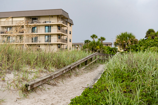 This is a photograph of a sandy beach pathway through a sand dune by the exterior of an older residential building in Cocoa Beach, Florida on a summer day.