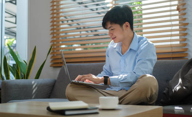 istock Smiling asian man sitting on couch and working online on laptop. 1480703623