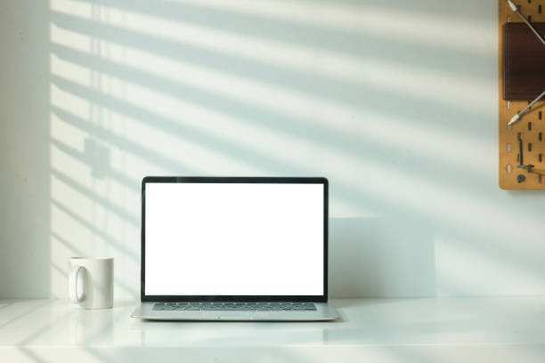 istock Front view of laptop computer and coffee cup on table with shadow from the jalousie on a white wall. 1480708577