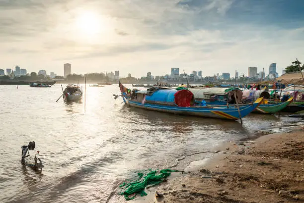 Small boats sit on muddy eastern bank of the river,much less developed than the riverside to the west.Setting sun reflecting on water,making silhouettes of small craft and distant city buildings.