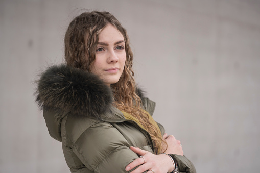 A close-up portrait of a confident looking young woman with a warm green winter jacket against a gray background. One person.