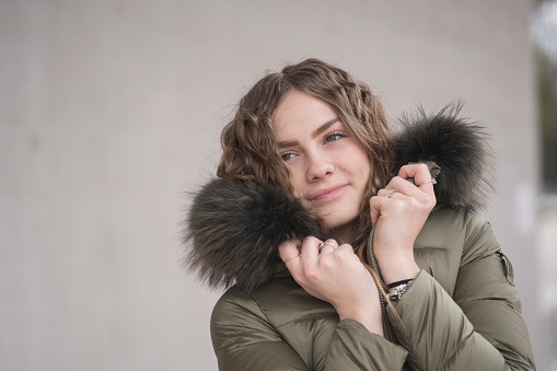 A close-up portrait of a confident looking young woman with a warm green winter jacket against a gray background. One person.