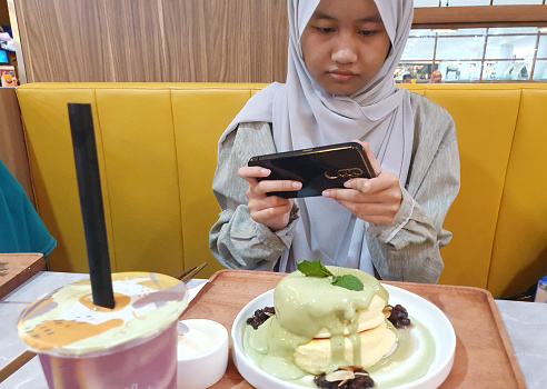 Samarinda, Indonesia. January 23th 2023. Happy Asian muslim teenage girl sitting in cafe and taking picture of her food with smart phone before eat