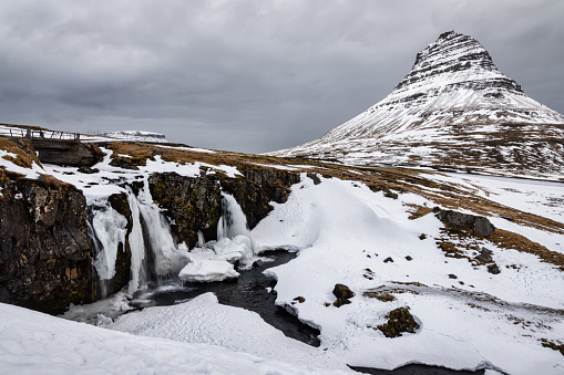 Kirkjufell Waterfall in Winter. Snow covered Kirkjufell Mountain and Kirkjufellsfoss - Kirjufell Waterfall in Winter under moody cloudscape. Kirkjufell - Icelandic: Church mountain, is a 463 m high mountain on the north coast of Iceland's Snaefellsnes Peninsula,near the town of Grundarfjordur. Kirkjufellsfoss Waterfall and Kirkjufell in Grundarfjörður, Snæfellsnes, Vesturland, Iceland, Nordic Countries, Northern Europe.