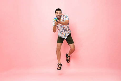 Handsome smiling young Caucasian tourist man playing with water gun and jumping in studio pink background for Songkran festival concept in Thailand and southeast Asia