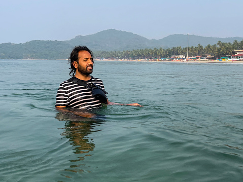 Stock photo showing Indian man chest deep in the water of the Indian Ocean, Palolem beach, Goa, India. The man is wearing a striped t-shirt and has a waterproof bag strapped across his chest to carry mobile phone and valuables.