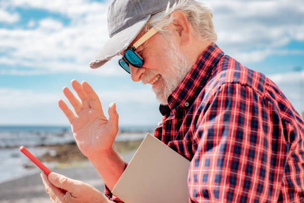 istock Smiling Senior Man Wave to Camera Having Video Call on Mobile Phone Walking Along the Beach. Elderly Pensioner with Red Checkered Shirt and Sunglasses Using Mobile Phone in Video Chat 1480746110