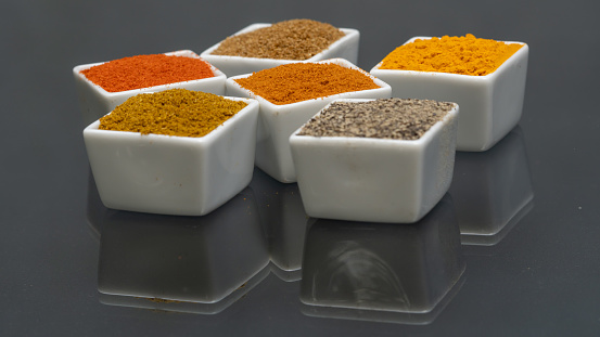 Colorful spices in white square bowls on black background, close up