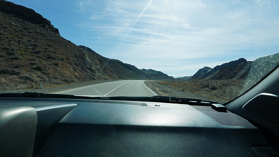 View of the road through the windshield. Cars are passing by. There are high rocky hills around the edges. Everything is covered with small dry bushes. There are dry trees and poles. White clouds