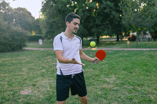 A young man is standing in a public park and preparing to start a pickleball game holding a racket.