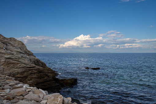Blue color of the sea with small waves. Blue sky with white clouds. Coastal rocks in the foreground. Limenas Thasou, capital of the island Thassos (Tassos), Greece.