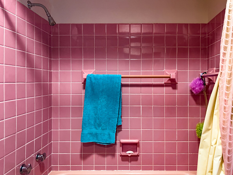 An outdated vintage bathroom highlighted in pink. Shower curtain and loofahs are visible.  An aqua colored towel hangs as an accent. The walls are tile squares.  From a middle-class home.