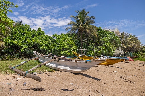 Galle, Sri Lanka - February 11th 2023:  Typical Sri Lankan fishing canoe made of fiberglass with a outrigger laying on the beach between garbage outside the Galle fish market