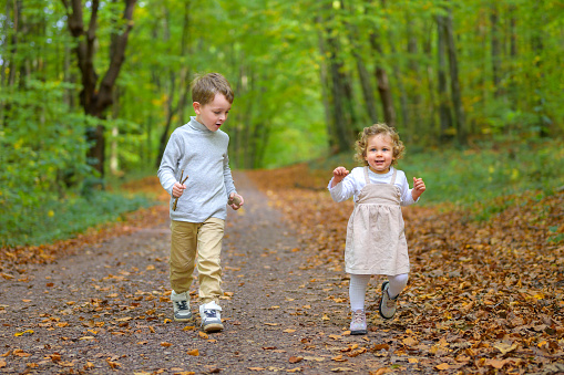 Brother runs happily with his little sister through the forest in autumn