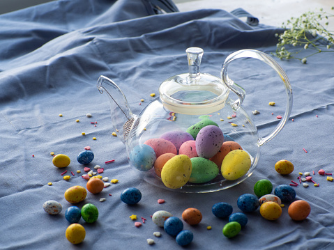 Composition with easter eggs in glass teapot surrounded by small chocolate eggs and sweet sprinkles. Easter tea concept. Tea pot full of colorful eggs on a blue background with copy space.