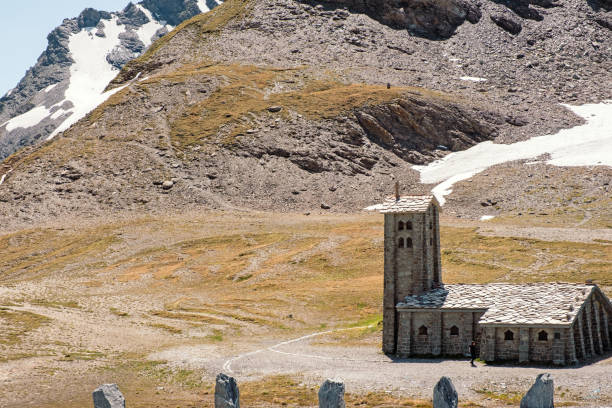 Col de l'Iseran, France - June 11, 2022:  Old stone chapel near de Col de l'Iseran, a famous mountain pass in the French Alps, early spring, snow covered extreme landscape with one female tourist hiking.