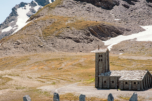 Col de l'Iseran, France - June 11, 2022:  Old stone chapel near de Col de l'Iseran, a famous mountain pass in the French Alps, early spring, snow covered extreme landscape with one female tourist hiking.