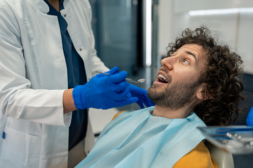 Hand of unrecognizable female dentist holding dental instrument near patient's mouth. Handsome young man with curly hair in dentist's chair looking at his dental specialist with trust.