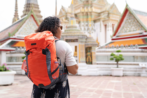 Temples are tourist attractions that calm the mind and feel relaxation. Portrait of African American backpack woman in causal with orange bag standing in the temple. Holiday vacation trip during weekend activity. Sightseeing at temple and see architecture