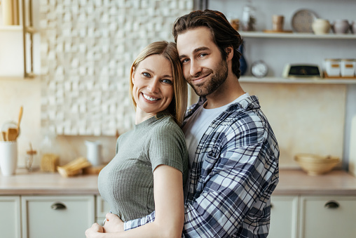 Smiling handsome european millennial guy with stubble hugs blond lady and enjoy tender moment at home at free time in light kitchen interior. Love and romance, family and people due covid-19 outbreak