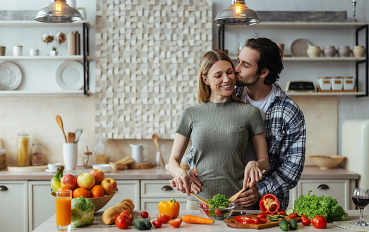 Smiling european young guy with stubble hugs, kisses blonde female, lady preparing salad at table with organic vegetables in kitchen interior. Cooking dinner at home, love, relationship due covid-19
