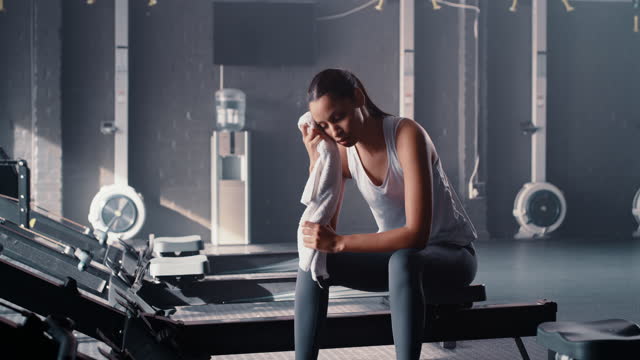 Woman, towel and sweating in gym for exercise, workout rest and training at rowing machine. Female sports athlete, sweaty and break in fitness club for recovery, endurance challenge and performance