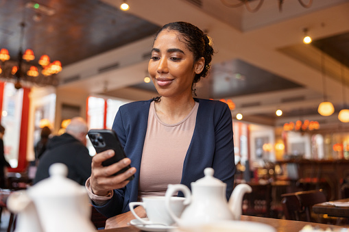 A dark-skinned female is smiling slightly while holding her cellphone and checking something on it. She has a kettle and some cups over the table where she is sitting.