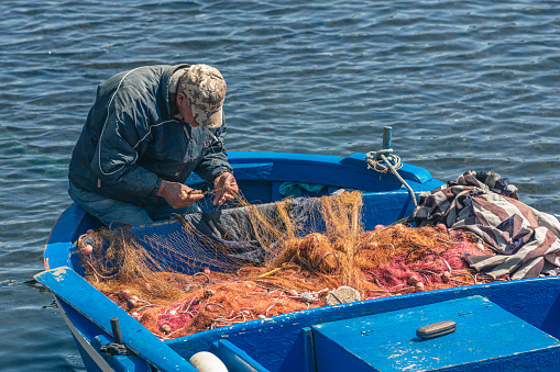 Bari, Puglia, Italy - April 8 2023: Fisherman with a hat repairing with a big needle a fishing net on an old blue wooden fishing boat at the port quay in Bari, Puglia Italy