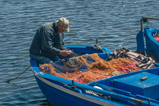 Bari, Puglia, Italy - April 8 2023: Fisherman with a hat repairing with a big needle a fishing net on an old blue wooden fishing boat at the port quay in Bari, Puglia Italy