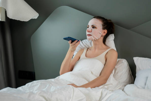 une femme occupée assise sur le lit de la chambre avec un masque cosmétique sur le visage, enregistre un message vocal sur son smartphone, prend des notes. soins de beauté du visage. - moisturizing cream audio photos et images de collection