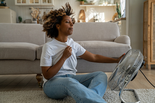 Summer heat. Young African American woman cooling down by ventilator at home, feeling unwell with high temperature during hot weather, sitting on floor in front of electric fan during extreme heatwave