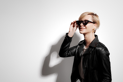 Confident young female in trendy black leather jacket and sunglasses standing against gray background in light with shadow and looking at camera