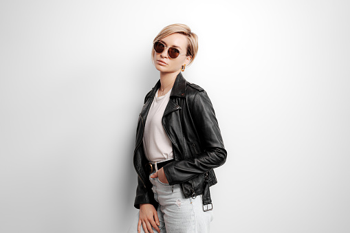 Confident young female in trendy black leather jacket and sunglasses standing against gray background and looking at camera