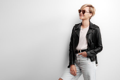 Confident young female in trendy black leather jacket and sunglasses standing against gray background