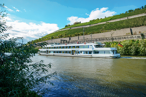 Recreational boat in Mid September with small quantity  of passengers and vineyards  on hillsides in Wurzburg.   Ship with German flag and blurred motion people  on it.\nElectrical wires of train : good  ecological trains.