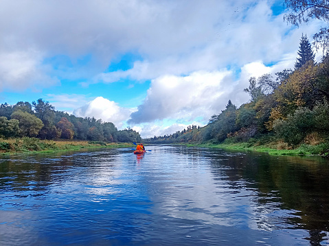 A group of people, in pairs in life jackets, are rafting on a calm river in orange inflatable kayaks in autumn