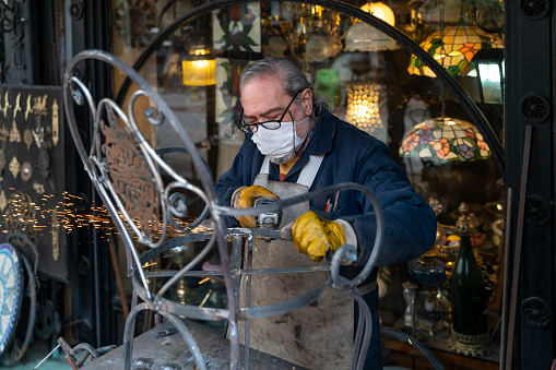 old blacksmith photographed working. he has a protective mask in his mouth. master carefully welds. Shot in natural light with a full-frame camera.
