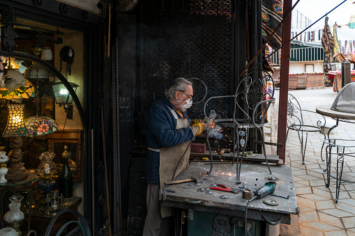 old blacksmith photographed working. he has a protective mask in his mouth. master carefully welds. Shot in natural light with a full-frame camera.